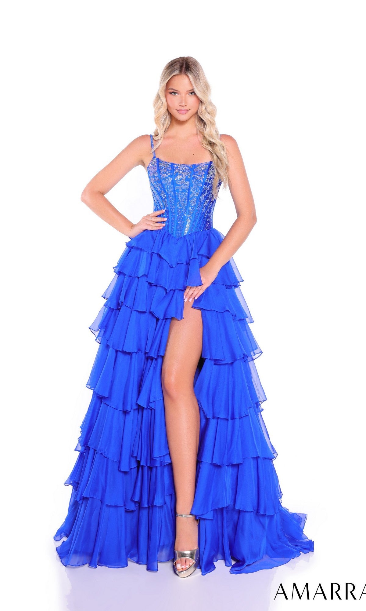 A woman stands against a white background wearing the Amarra 89459 Lace-Up Long Tiered Prom Dress in bright blue, featuring a high slit and an embellished corset-style bodice.